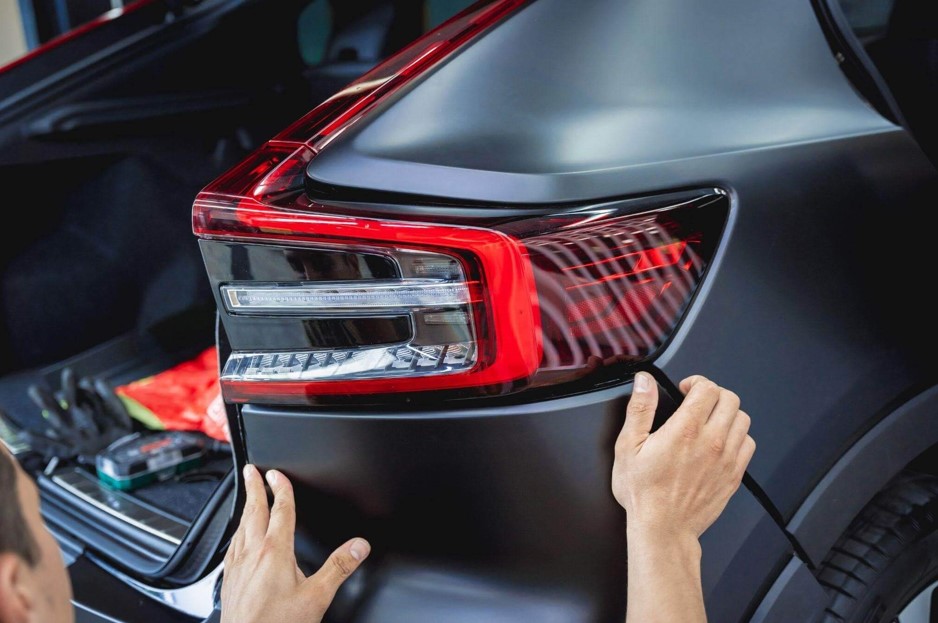 Um homem (visto parcialmente no canto inferior esquerdo) está com as mãos posicionadas sobre o para-choque traseiro, logo abaixo da lanterna, como se estivesse ajustando, encaixando ou inspecionando a peça.