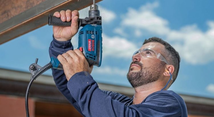 Um homem com óculos de proteção e protetores auriculares opera uma Furadeira de Impacto Bosch GSB13 RE azul. Ele utiliza as duas mãos para perfurar uma viga metálica acima de sua cabeça contra um céu azul com nuvens. A imagem destaca o uso profissional e ergonômico da ferramenta em ambiente externo.