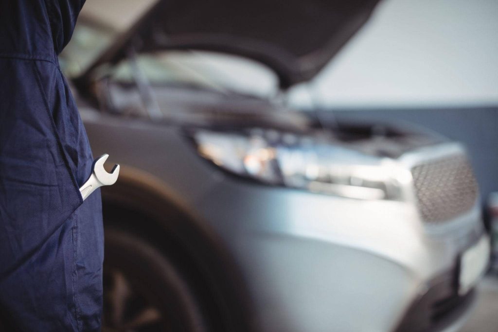 Close-up de um mecânico com uma chave de boca no bolso do uniforme, observando um carro prata com o capô aberto ao fundo; uma cena que remete à busca por sinais de embreagem falhando durante uma revisão profissional.
