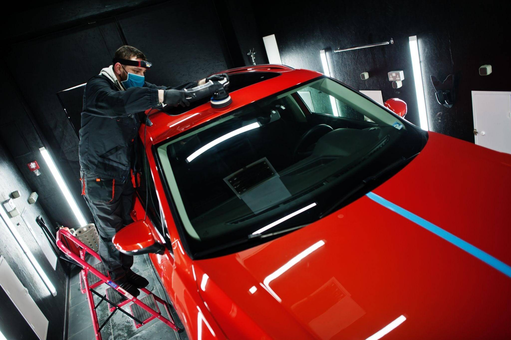 Profissional de estética automotiva profissional equipado com máscara e lanterna de cabeça realiza o polimento técnico no teto de um carro vermelho brilhante, utilizando uma politriz orbital em um estúdio de detalhamento com iluminação em LED.