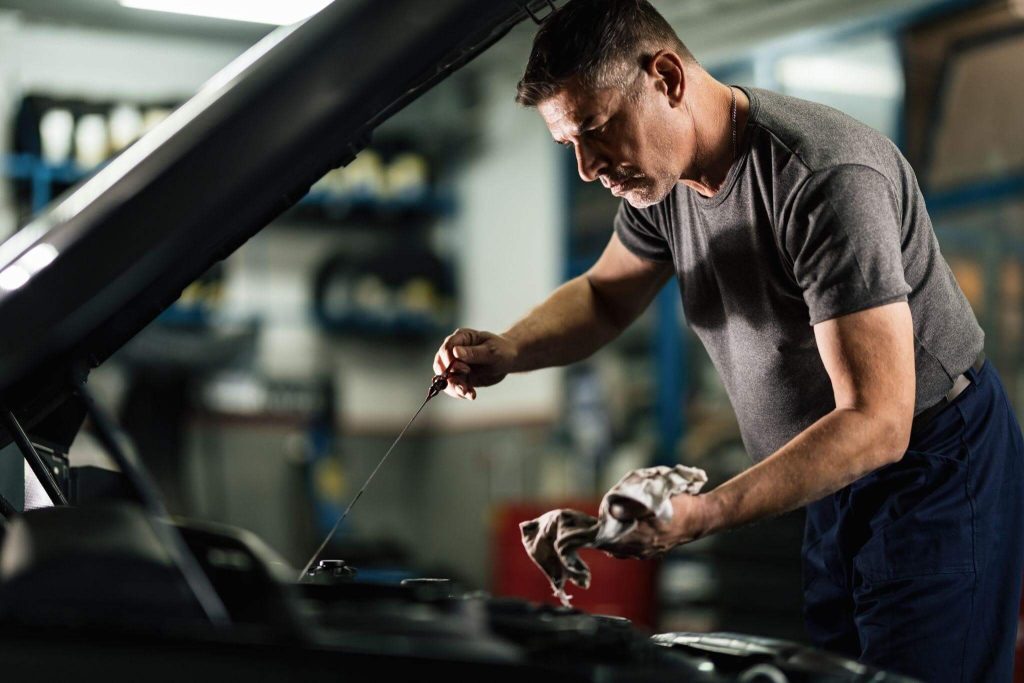 Homem em uma oficina mecânica com o capô do carro aberto, segurando a vareta de medição para verificar o nível e o estado do lubrificante. Ao fundo, prateleiras organizadas sugerem o que comprar quando você for fazer a troca de óleo do motor.